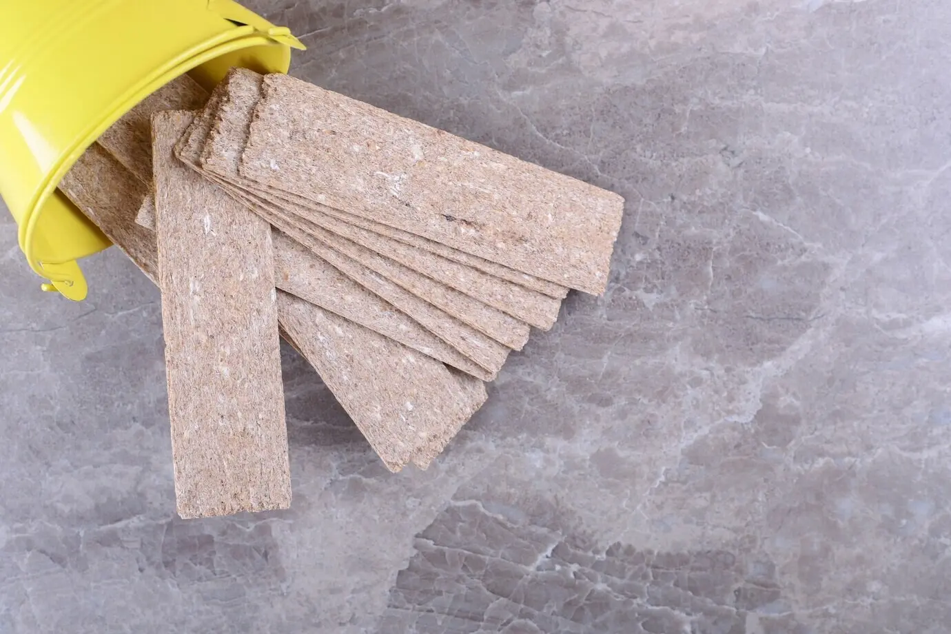 Crispbread falling from an overturned yellow bucket on the marble surface.